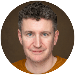 Man with short curly hair, wearing a brown jumper, against a plain brown background.