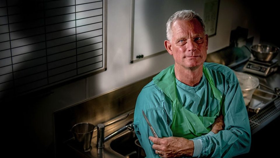 A man in green scrubs poses confidently with a surgical instrument in a clinical setting.