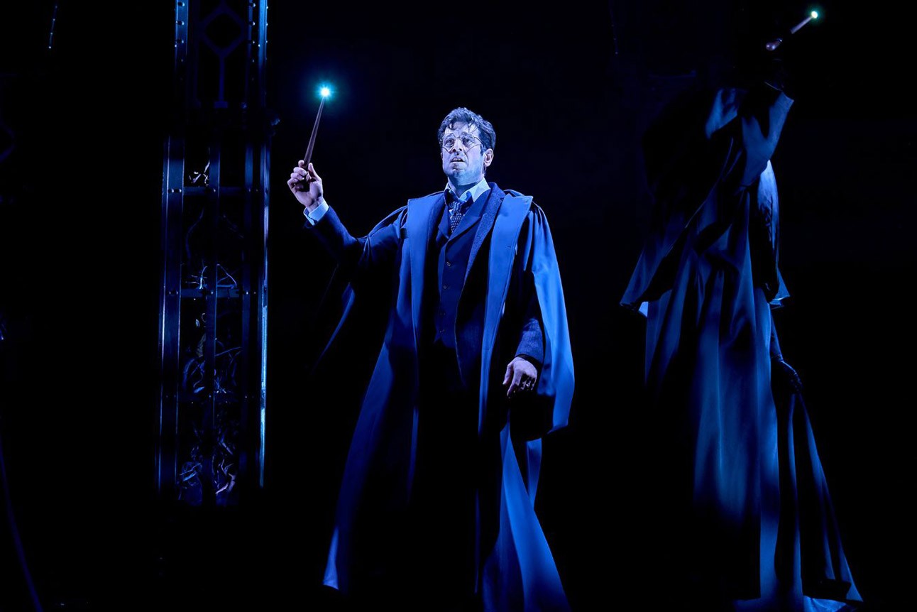 A person in a blue robe holds a glowing wand on a dark stage.
