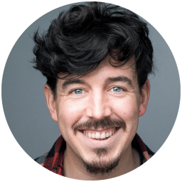 Smiling man with curly dark hair and a beard, wearing a checked shirt, grey background.