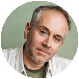 A man with short grey hair and a beard, wearing a light shirt, looks at the camera.