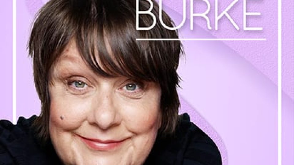 Kathy Burke poses with a warm smile, wearing a dark top on a purple backdrop.