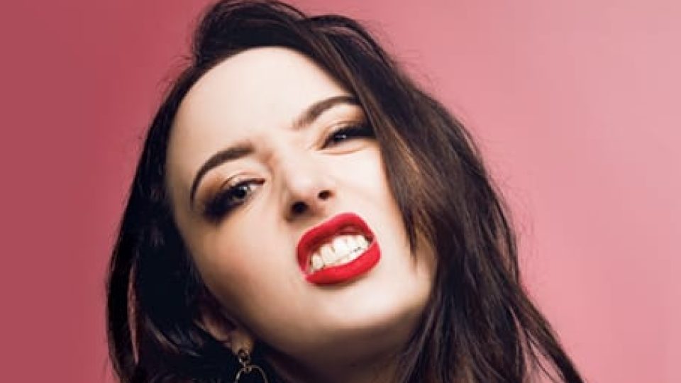 Fern Brady with long dark hair, wearing bold lipstick, against a pink background.
