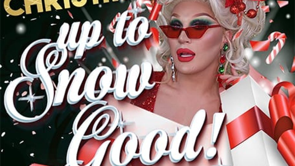 A drag performer in red attire, posing with festive decorations and a playful expression.