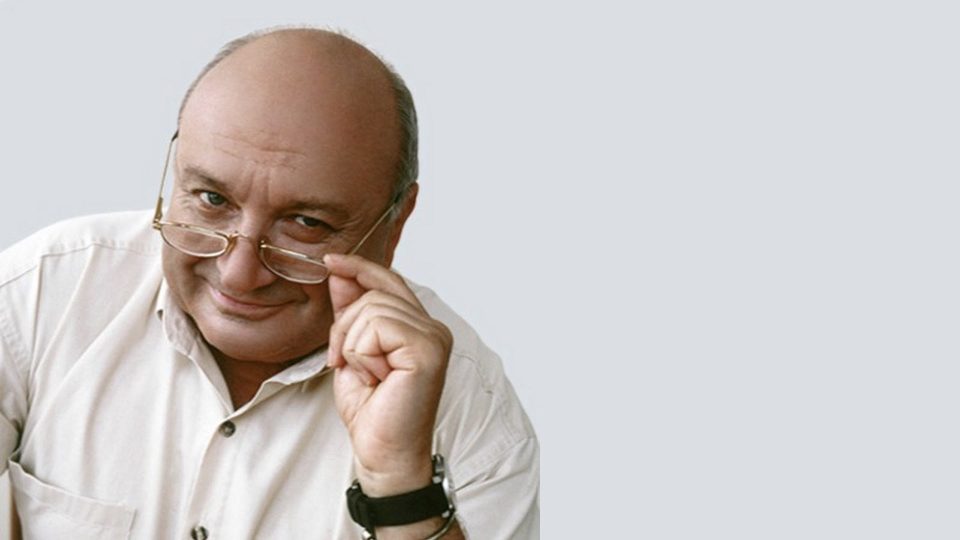 Mikhail Zhvanetsky smiles while adjusting his glasses, wearing a white shirt.