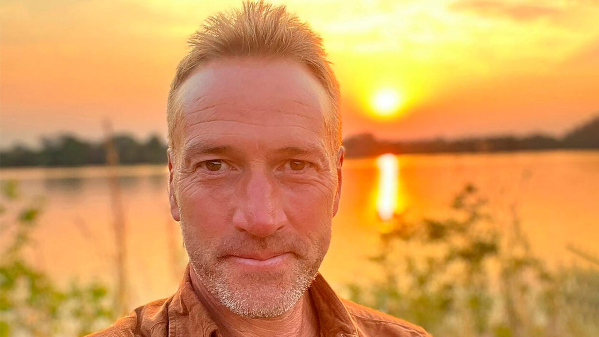 A man with short hair smiling at sunset, reflecting on water behind him.