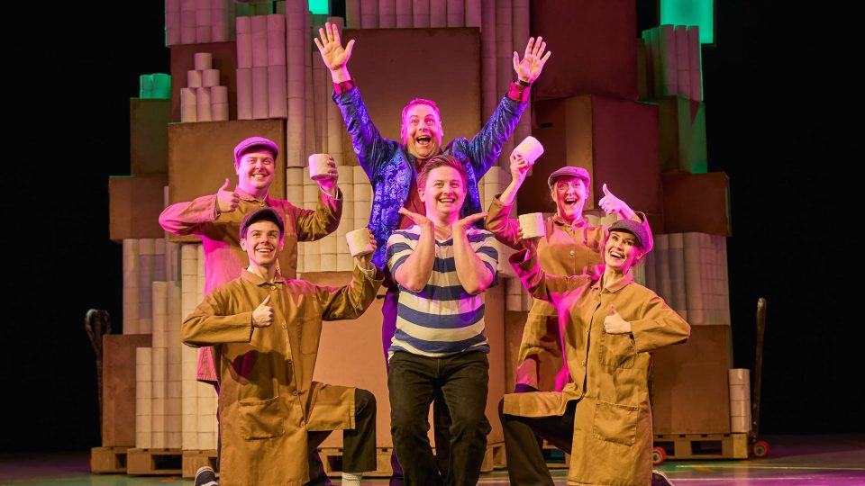 A group of performers in brown costumes cheerfully pose with cups on stage.