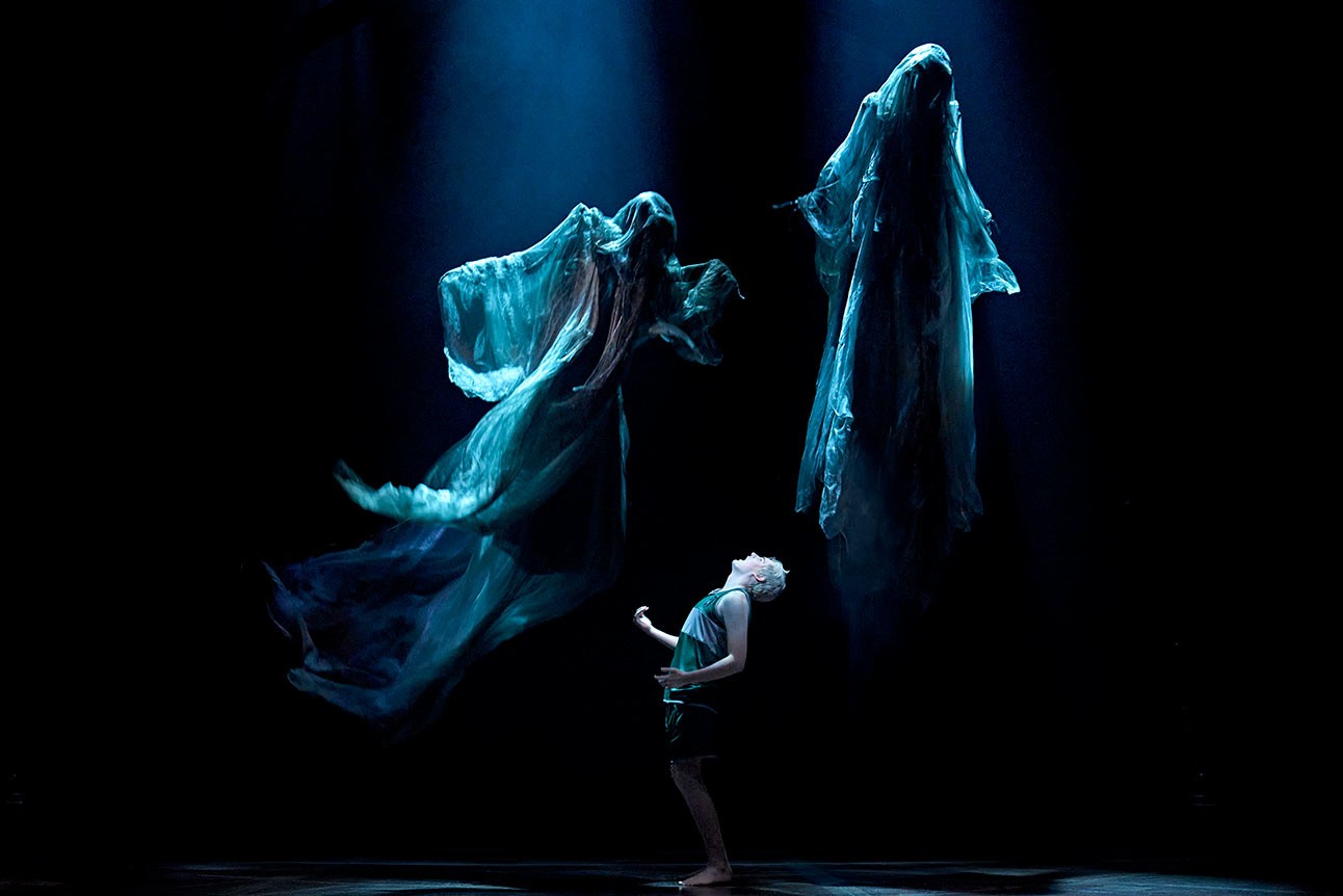 A performer in spotlight looks up at two ethereal, floating figures in blue-lit stage scene.