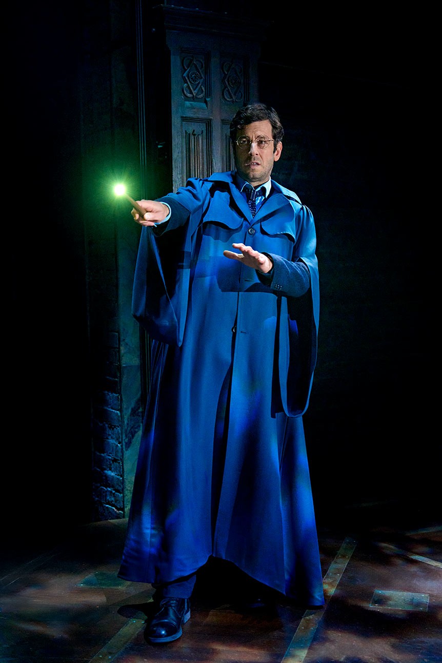 Actor in blue robe holds glowing green wand on dark stage, performing in "Harry Potter and the Cursed Child".