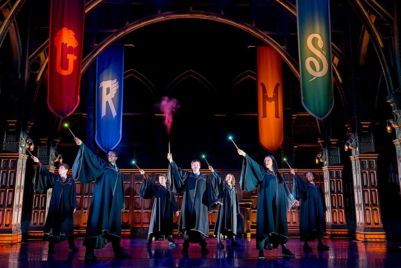 Actors in robes raise wands on stage, colourful house banners hang in the background.