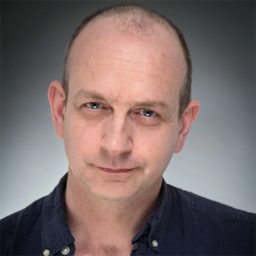 Bald man in a dark shirt against a grey background, looking directly at the camera.
