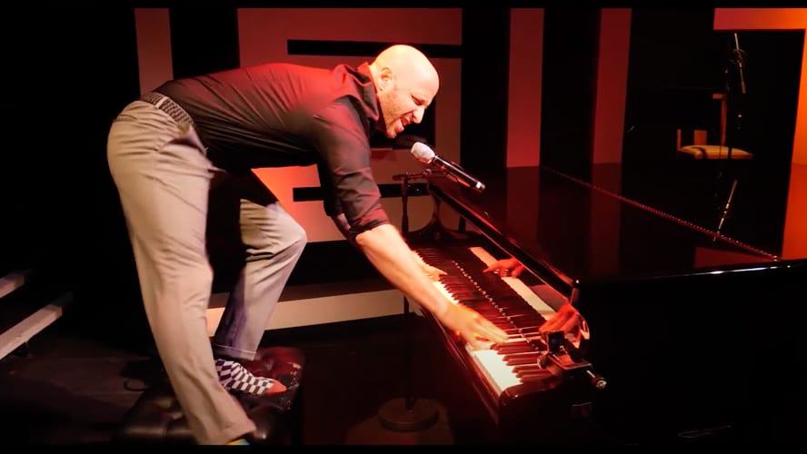 A man plays a grand piano, wearing a black shirt, under red lighting.