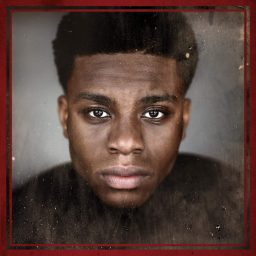 Femi Akinfolarin in costume, standing confidently against a dark background with subtle stage lighting.