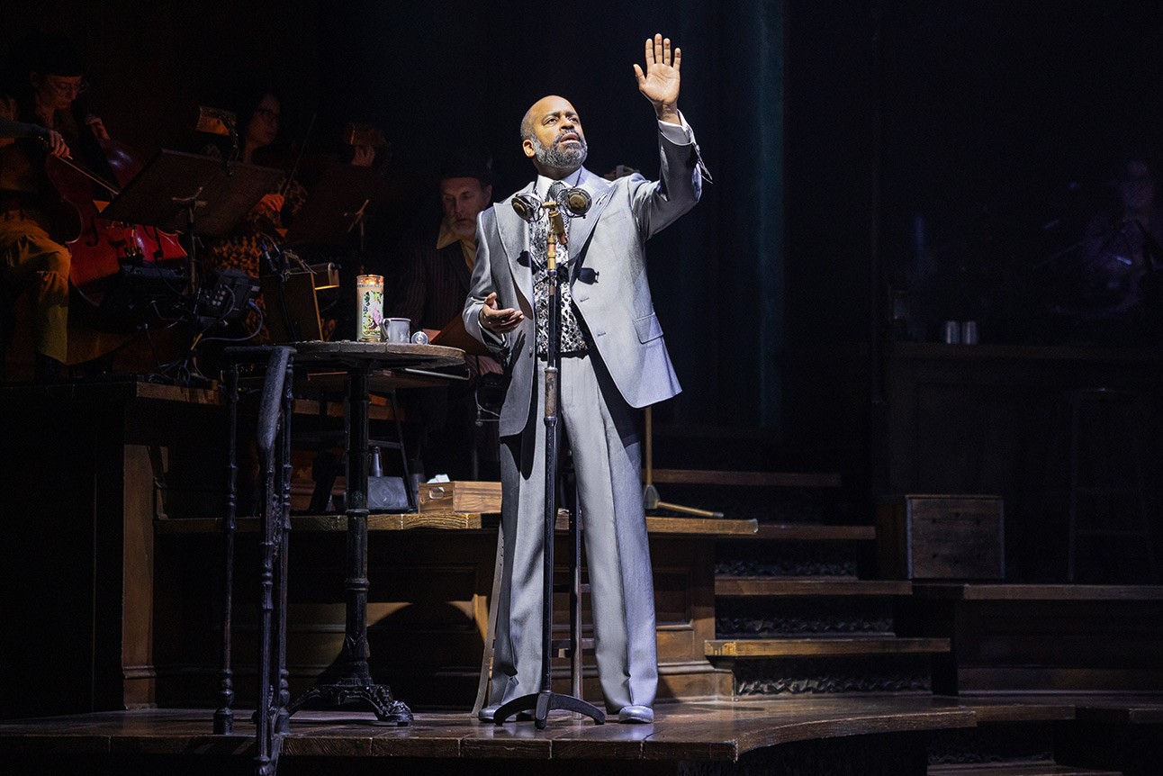 Daniel Breaker performs on stage in Hadestown, wearing dark costume against a dimly lit background.