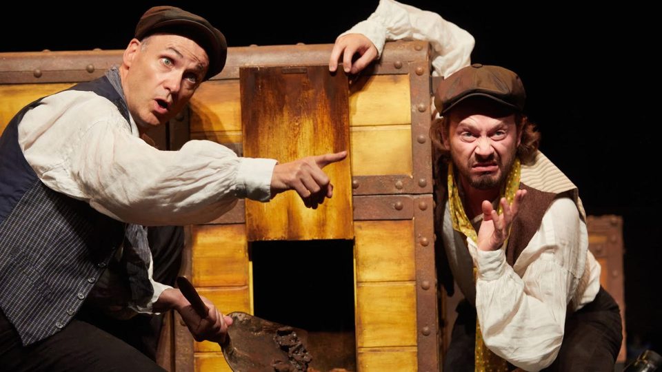 Two actors in historical costumes express surprise near a wooden chest.