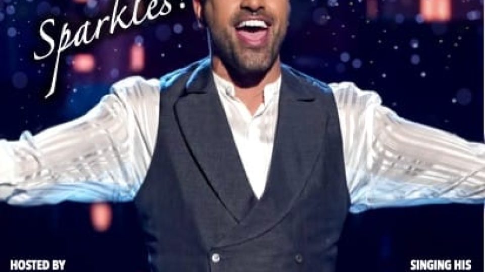 Dr Ranj, in a black suit, sings joyfully on stage with a sparkling background.