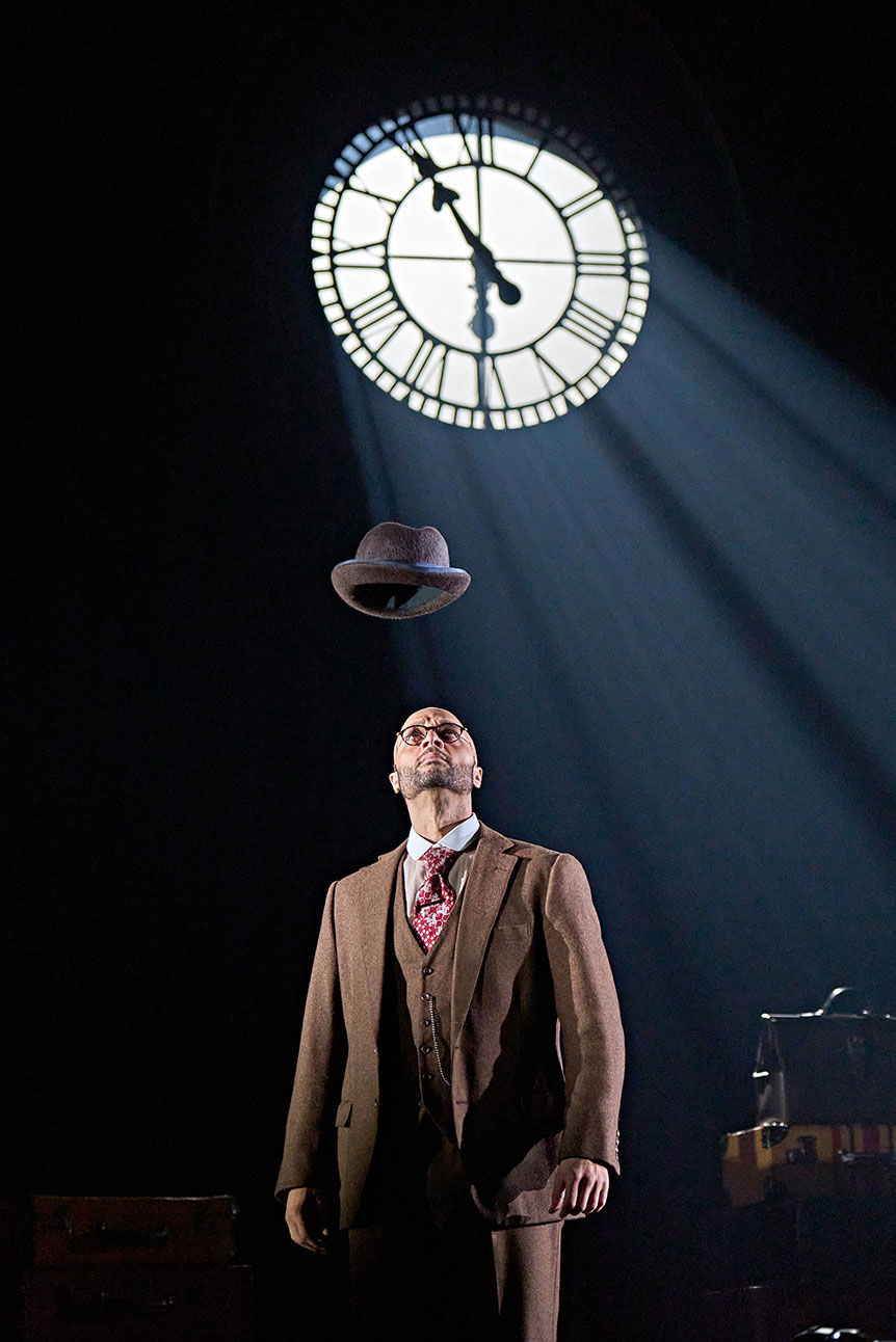 A man in a suit gazes upwards at a levitating hat, under a large clock face.