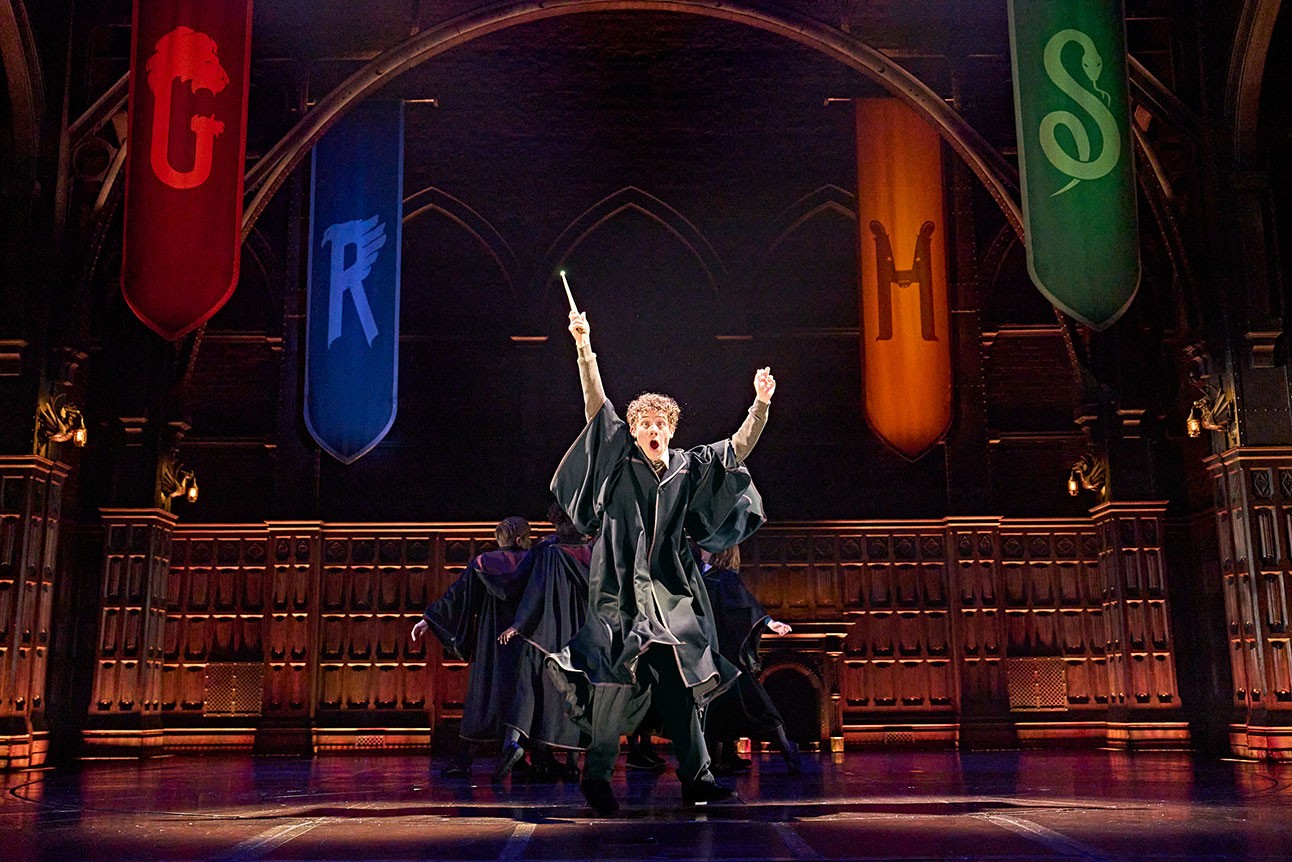 Actor on stage raises wand, surrounded by Hogwarts banners; blue, red, and green visible.