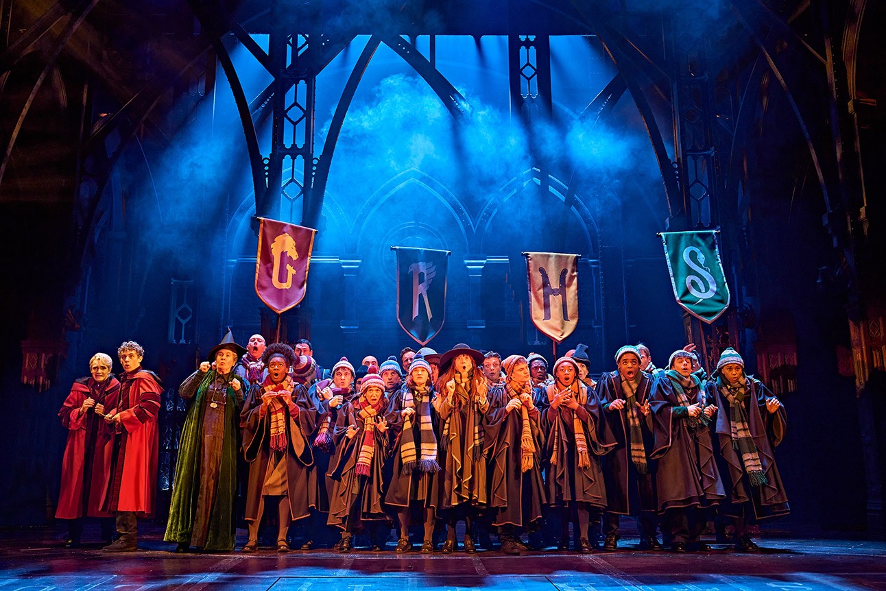Stage scene from "Harry Potter and the Cursed Child" with actors in Hogwarts robes under house banners.