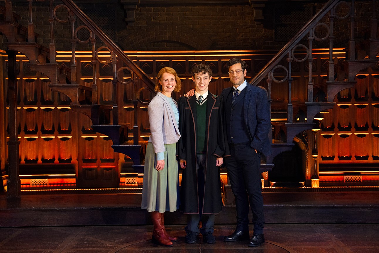 Three actors in costume stand on stage, with wooden stairs and warm lighting in the background.
