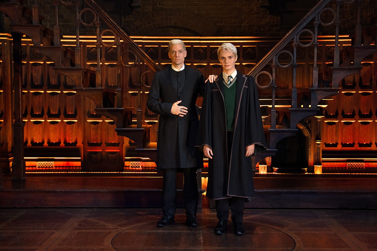 Two actors in dark robes stand on stage, wooden staircase backdrop, warm lighting.