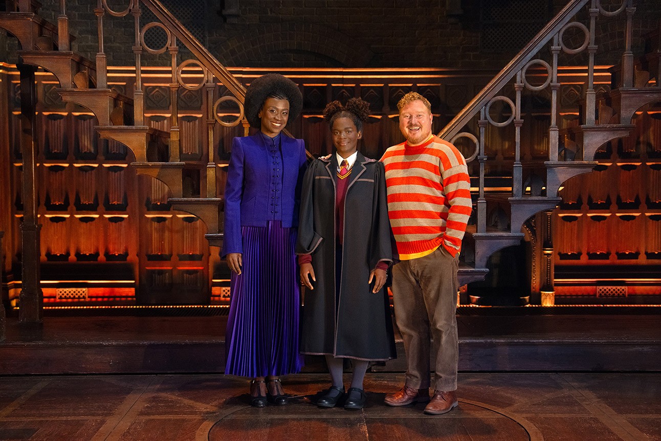 Three actors on stage, one in Hogwarts robe, others in colourful outfits, wooden backdrop.