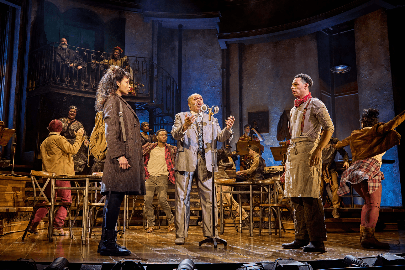 Hermes sings into a microphone while Orpheus and Eurydice stare at each other