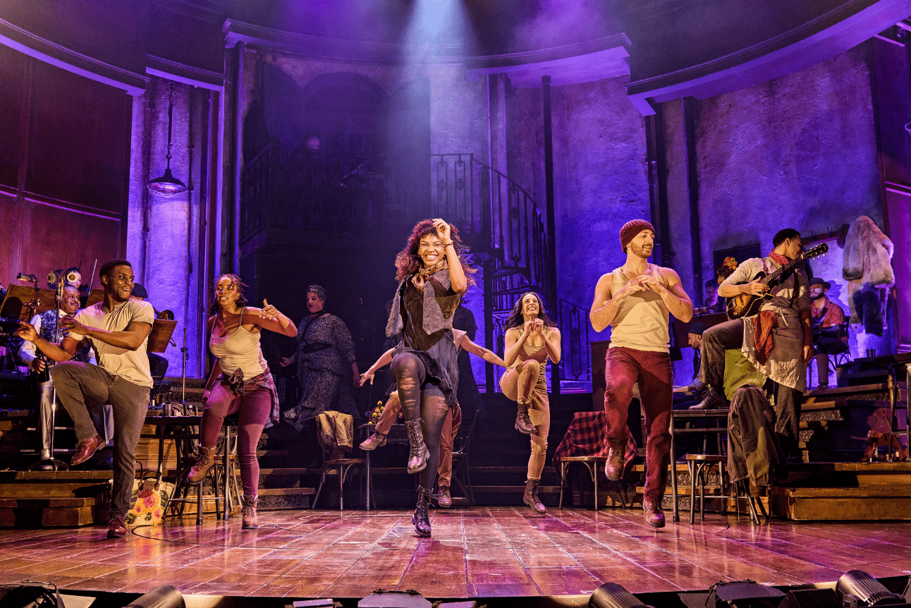 The cast of Hadestown dance enthusiastically. The stage is lit in a vibrant purple colour