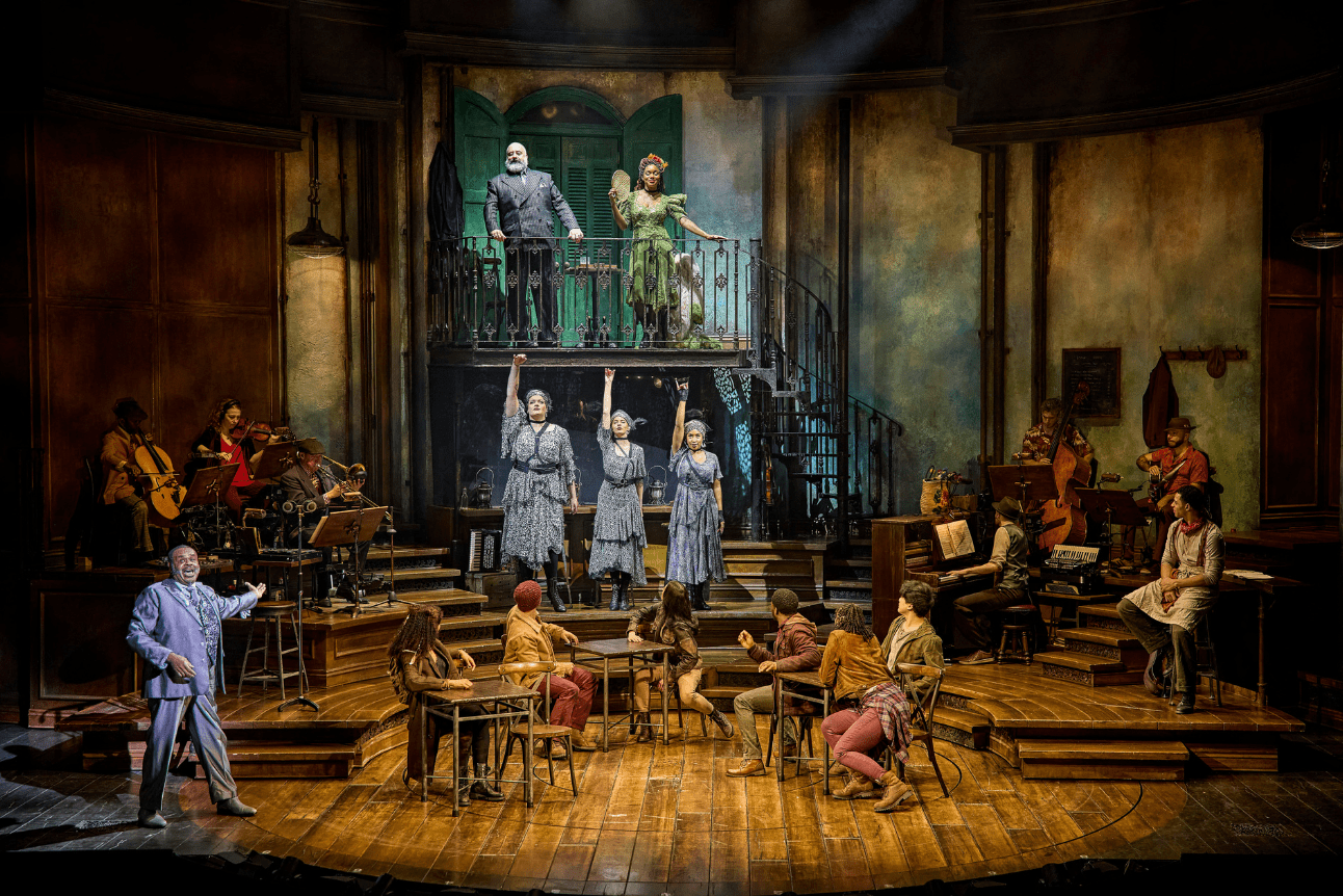 A wide shot of the cast of Hadestown performing on stage - There are people on the balcony, playing insturments, sat on chairs at the centre