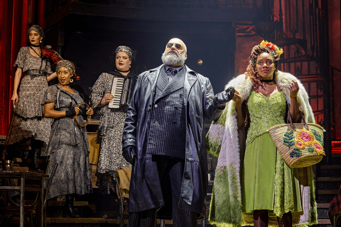 Hades and Persephone hold hands while the Fates watch on from behind