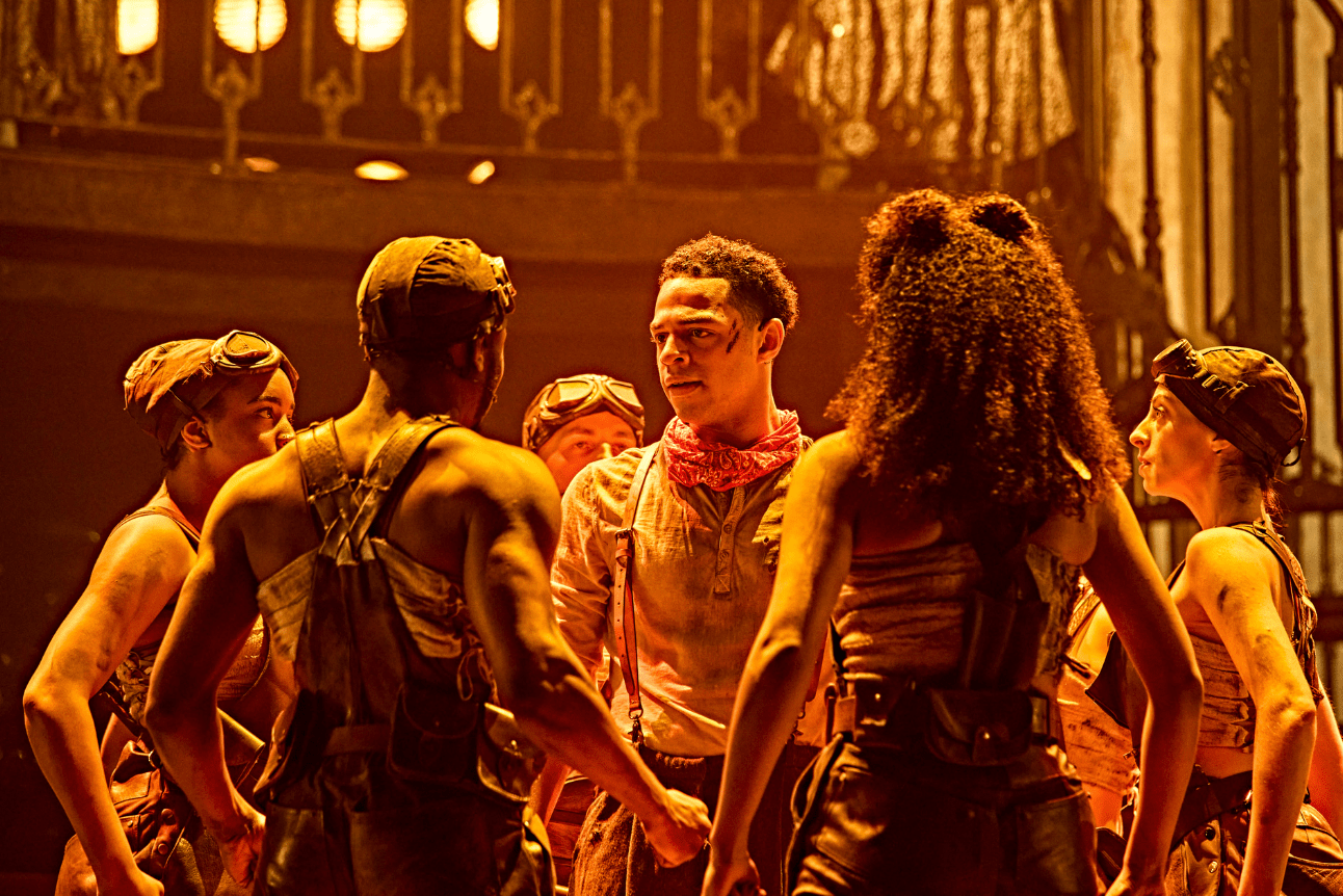 The workers in Hadestown surround Orpheus. They are bathed in a bright orange light