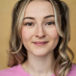 Young woman with blonde hair in pigtails, wearing a pink shirt, smiling against a yellow background.