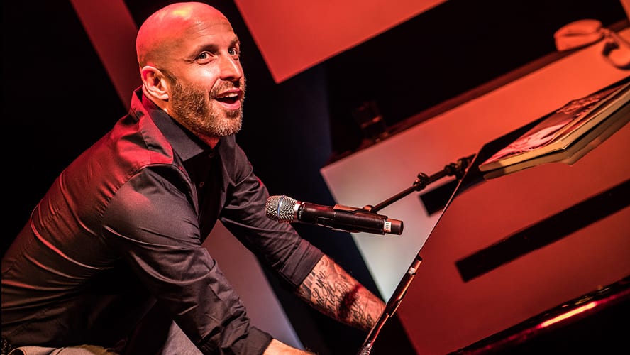 A man smiles while playing piano, with red lighting and a microphone nearby.