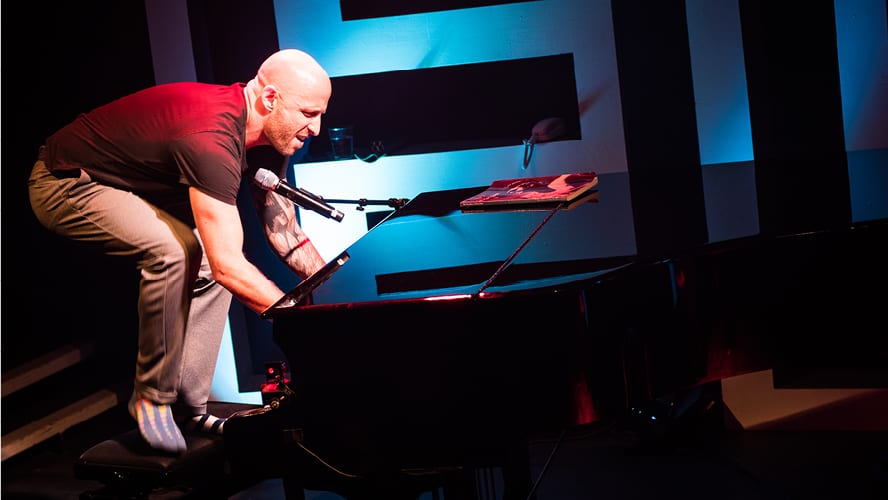A man energetically plays piano, wearing a black t-shirt and grey trousers, with blue accents.