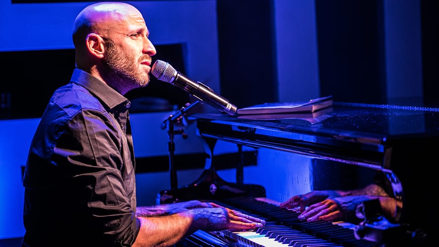 A man sings into a microphone while playing piano, illuminated by blue lighting.
