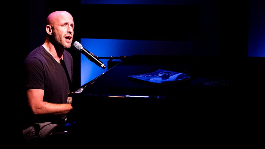 A man sings into a microphone while playing piano, illuminated by blue light.
