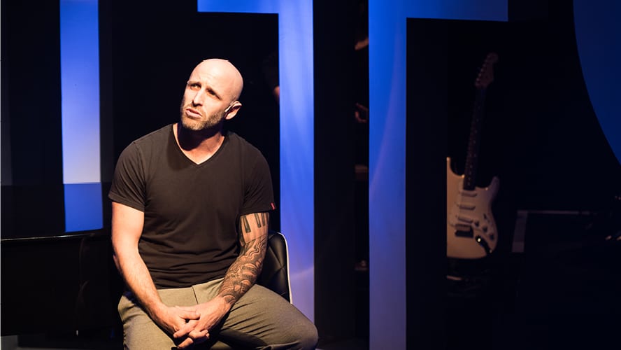 A man sits thoughtfully on a chair, wearing a black t-shirt, with blue lighting behind.