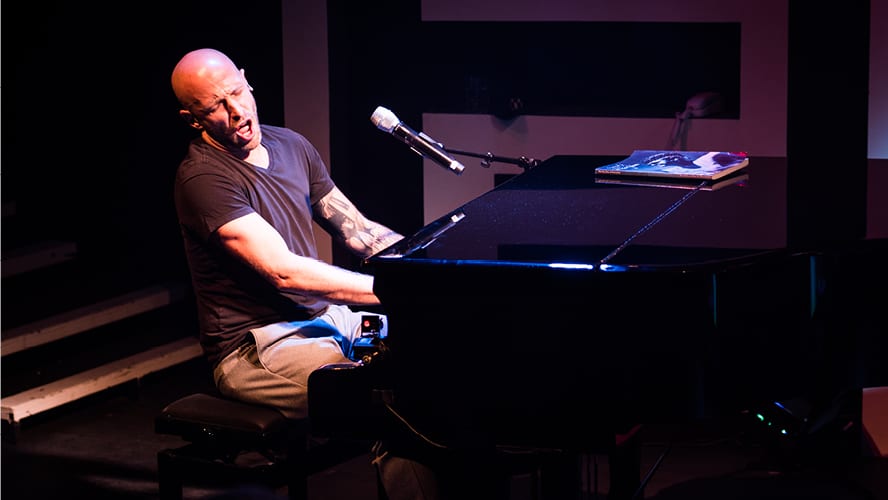 A man plays a black piano passionately, wearing a black t-shirt and grey trousers.