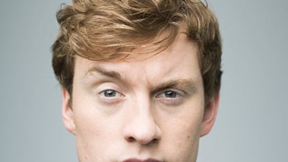James Acaster has tousled blond hair and a serious expression against a grey background.