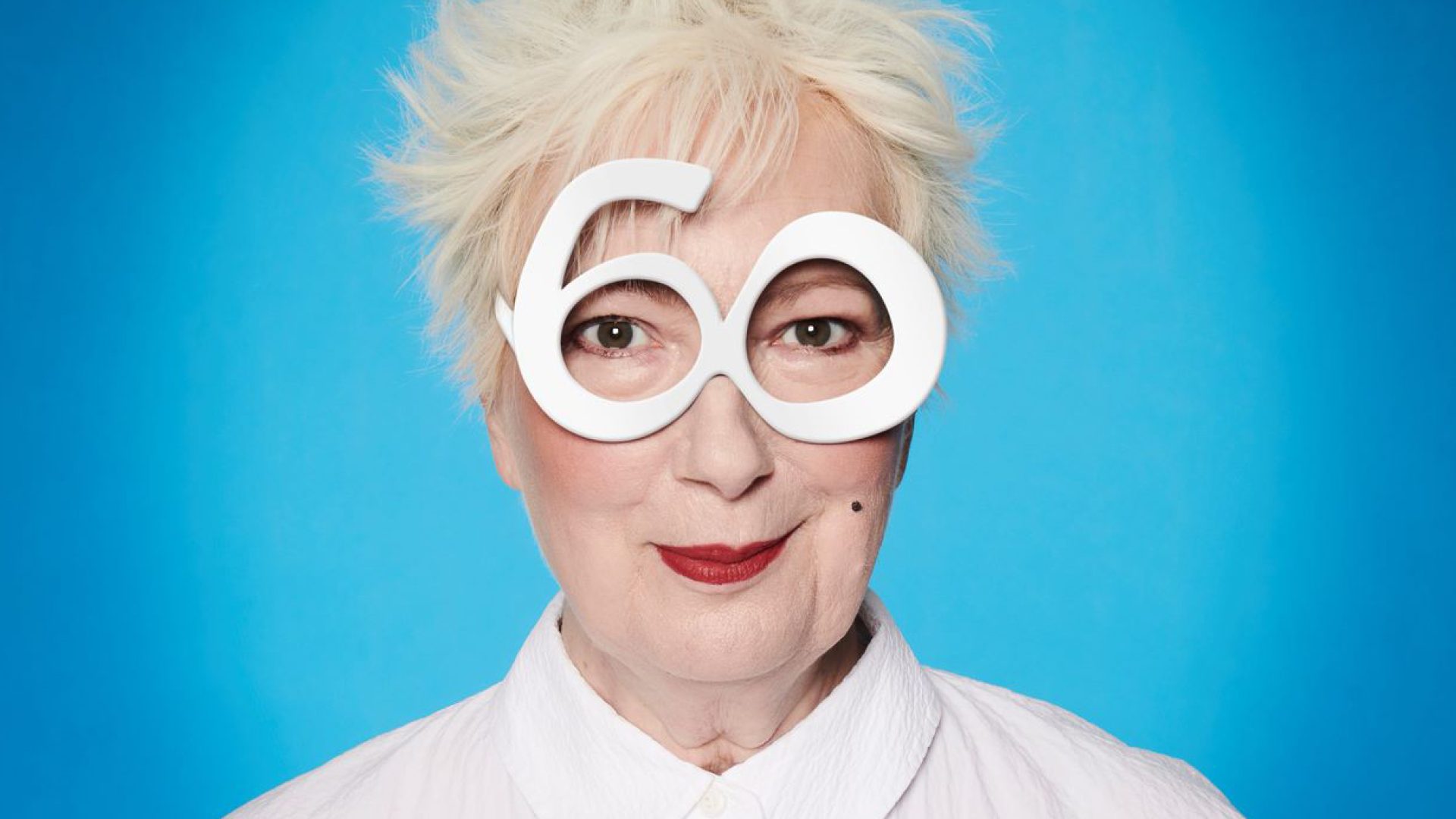 Jenny Eclair with white hair and large glasses shaped like the number 60.