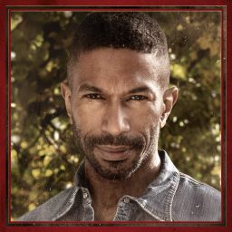 Juan Jackson swings a guitar mid-performance, wearing dark clothing against a dimly lit stage background.