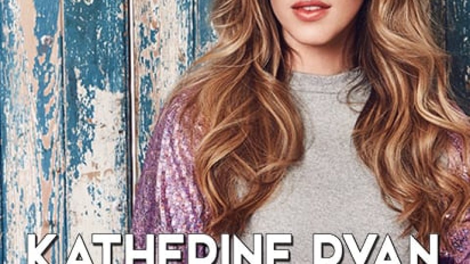 Katherine Ryan stands in a grey top with a sequined purple sleeve, blue wooden background.