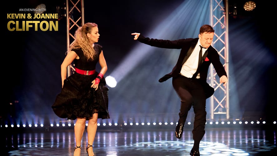A man in a tuxedo and a woman in a black dress dance on a polished stage.
