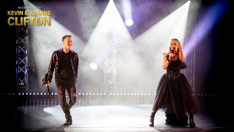 A man in a leather jacket and a woman in a black dress perform on stage.