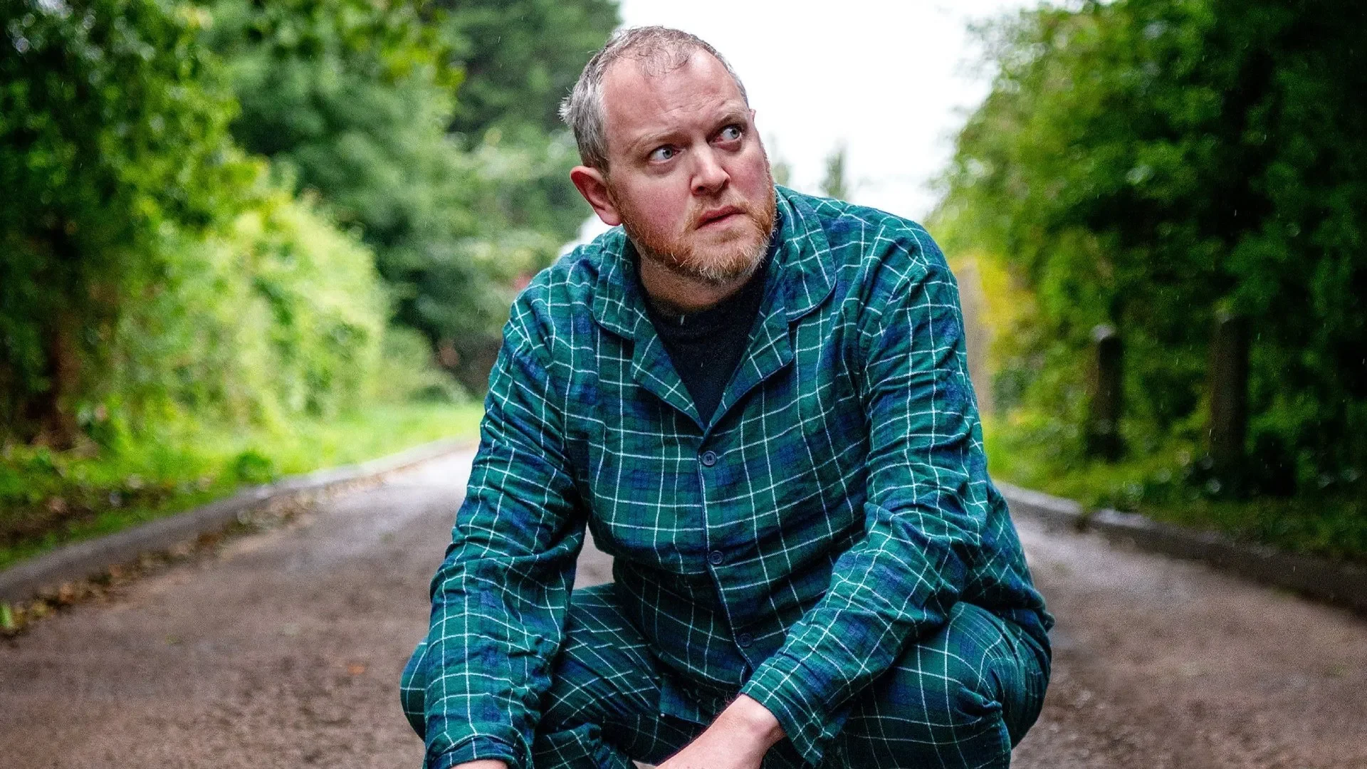 Miles Jupp squatting on a road, wearing a green checked pyjama set.