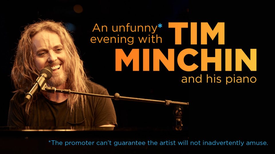 A man with long hair smiles while playing piano, illuminated by stage lights.