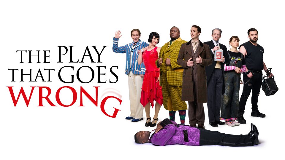 Six actors in colourful costumes pose comically; one lies on the floor in a purple jacket.
