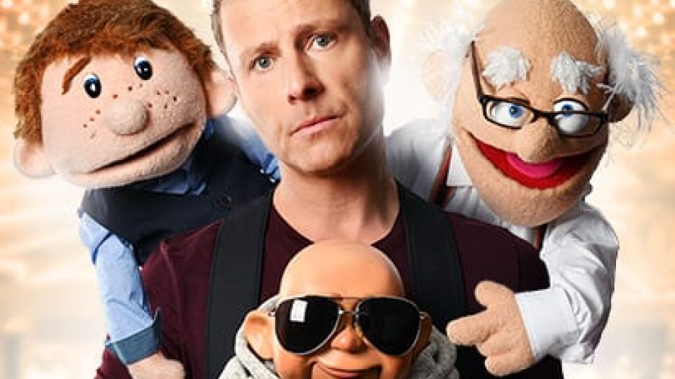 Paul Zerdin smiles with three colourful puppets against a softly lit theatre backdrop.
