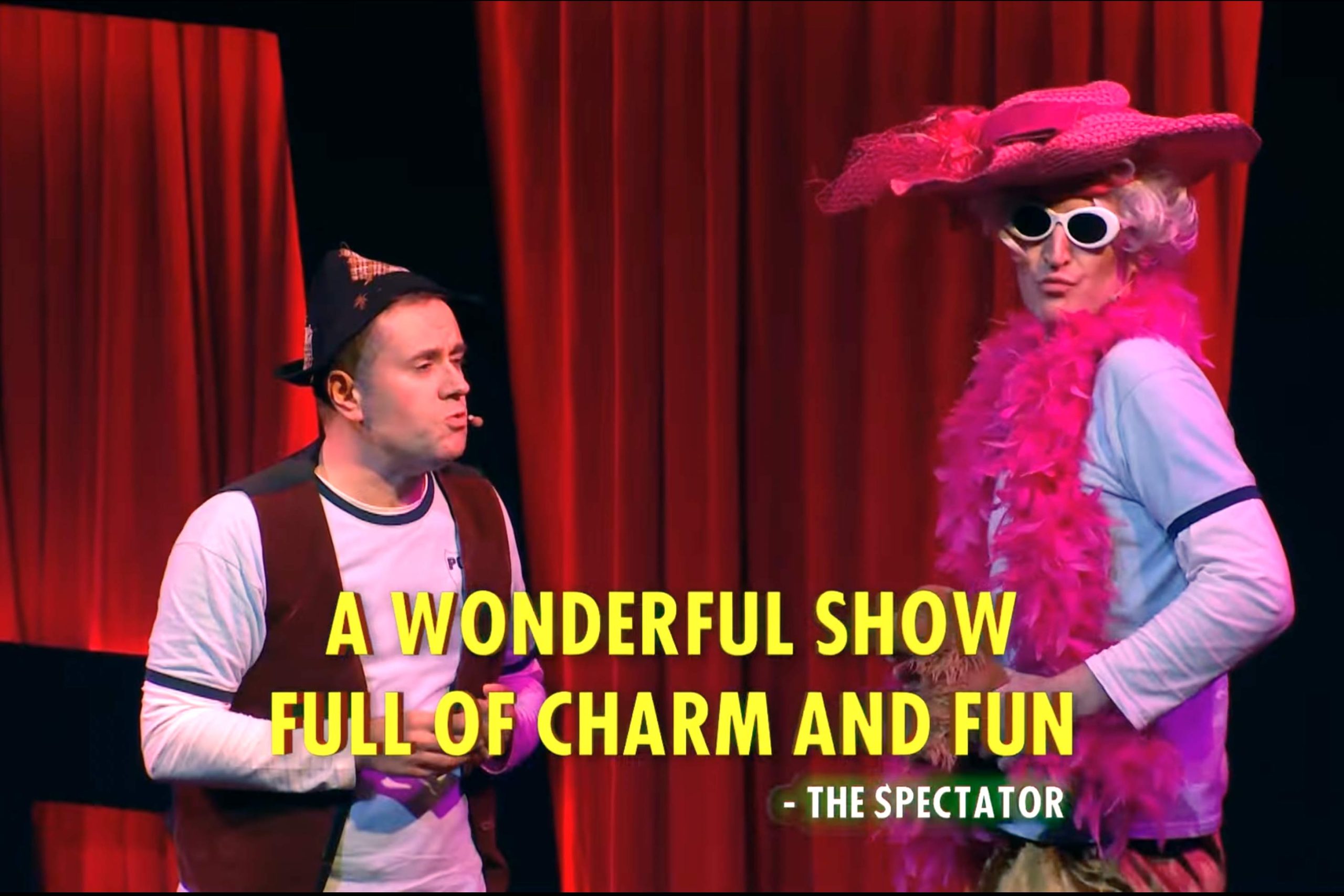 Two performers on stage; one in a pink feather boa and hat, text overlaying red curtains.