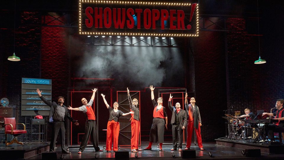 Seven performers in red and black costumes pose dramatically under stage lighting.